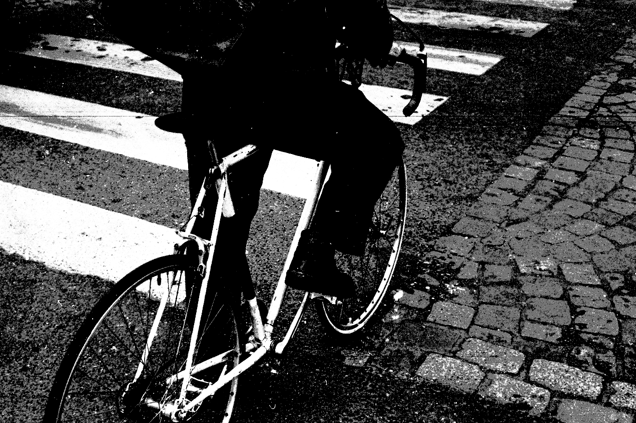 Photographie en noir et blanc, un homme sur une bicyclette longe un passage pour piéton, on ne voit pas son visage, l'image a été coupée.